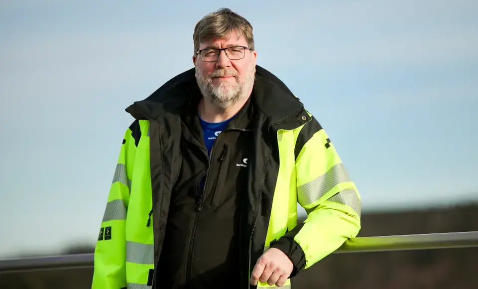 Man i gul signalväst och glasögon står framför ett räcke, yrkesklädd personal inom vägarbeten.
