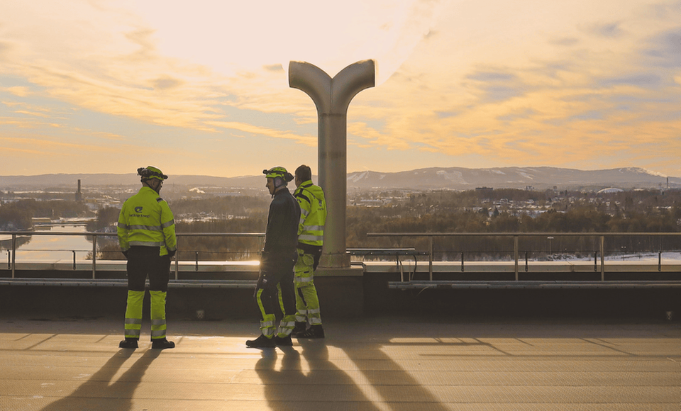 Två vägarbetare i varselkläder blickar ut över en vinterstad i solnedgången. Stora konstruktioner och ett snötäckt landskap syns i bakgrunden.